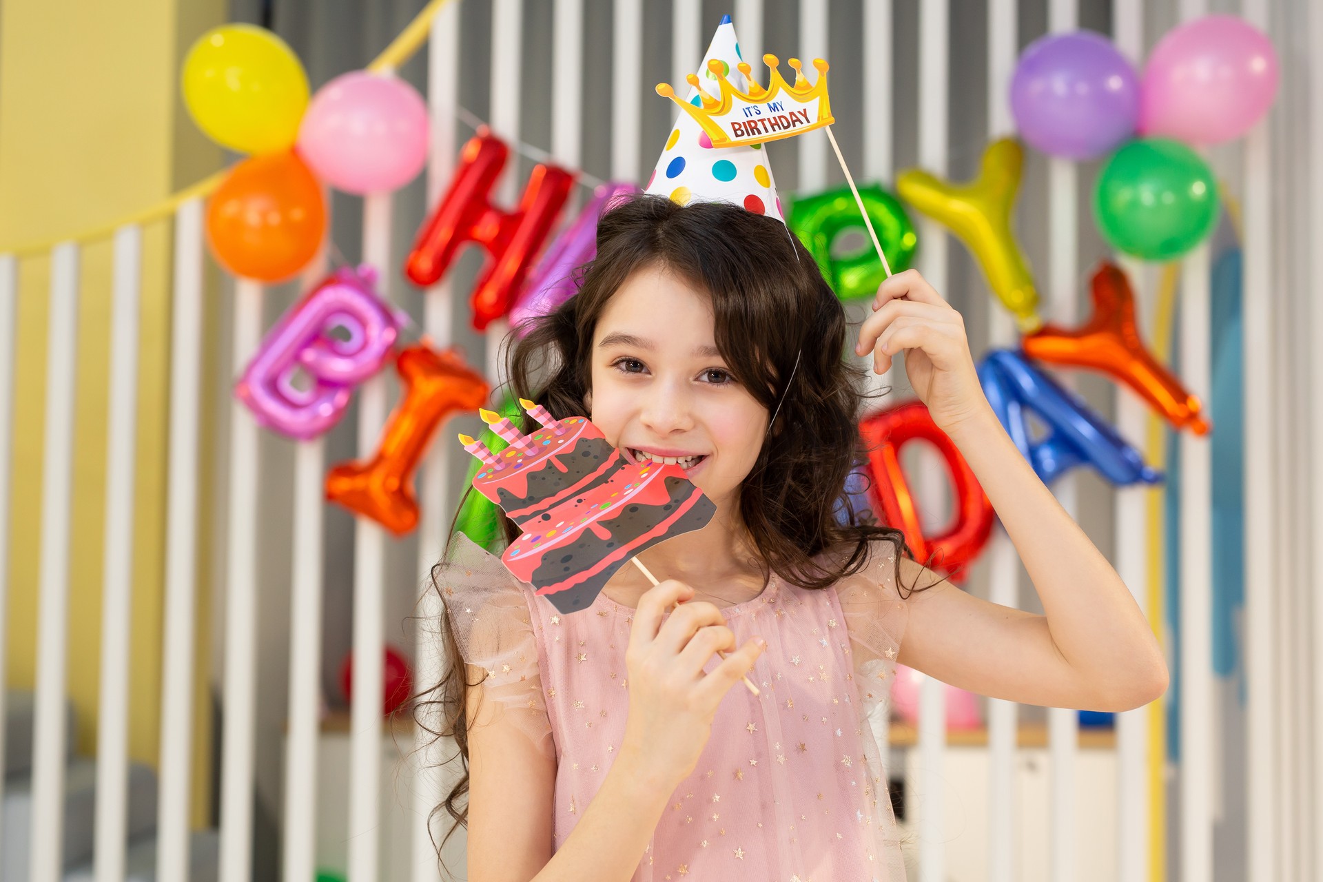 The birthday girl in a festive hat, with a pipe and props is having fun on the background of a multi-colored wall with balloons.